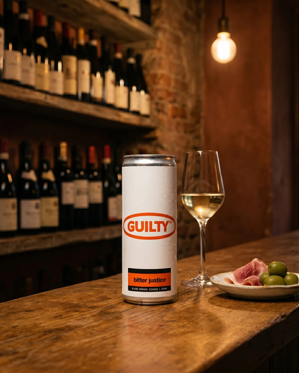 Bitter Justice 250ml can on a worn timber wine-bar counter, beside a glass of white wine and a small plate of prosciutto and green olives. Bottle shelves and exposed brick behind. Edison bulb overhead.