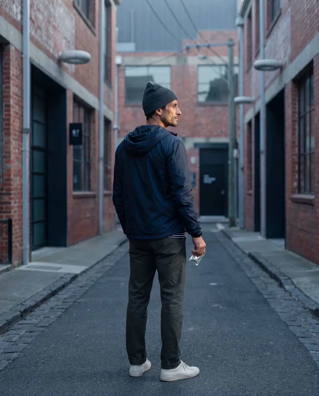 Subject MELB-01 standing in a Melbourne laneway between red brick warehouses. Navy hooded windbreaker, charcoal beanie, dark trousers, white sneakers. Holding a small clear sachet.