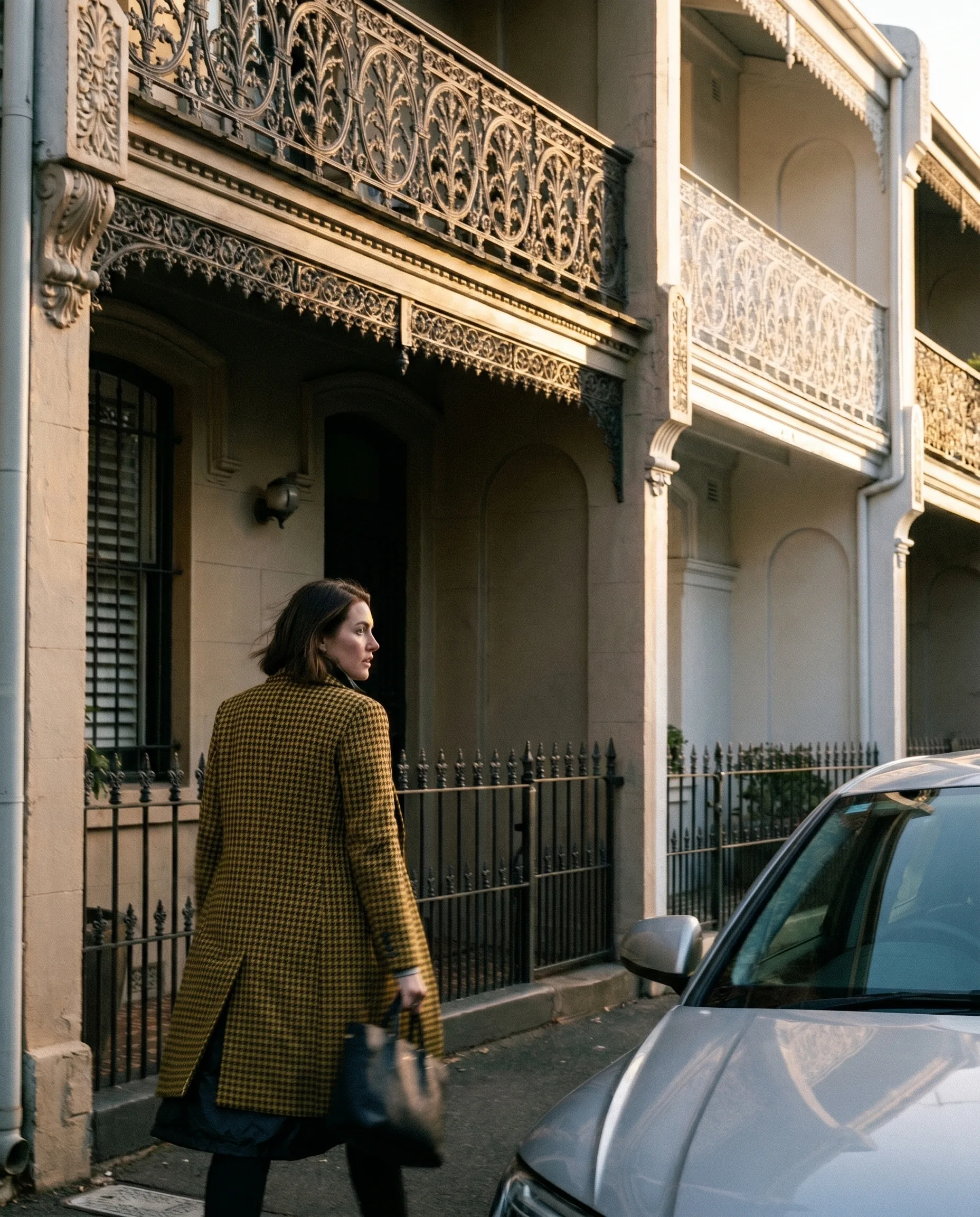 Subject SYD-02, mid-stride along a Sydney terrace street with cast-iron lacework balconies. Mustard-yellow houndstooth wool overcoat, black skirt, slim black leather handbag. Late afternoon golden hour, profile read.