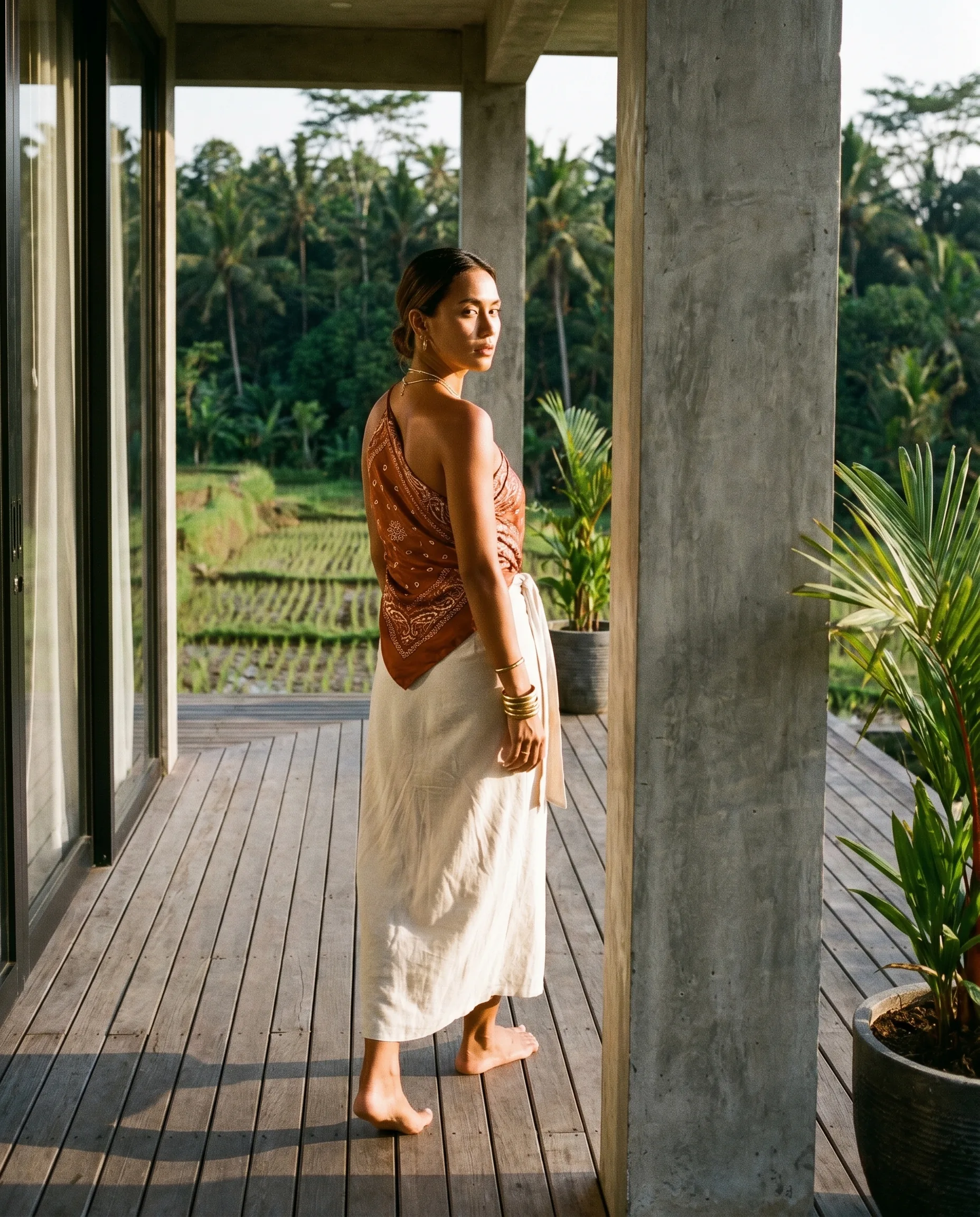 Subject BNE-03 on a Villa Soma deck at golden hour. Burnt orange paisley silk top, cream linen wrap skirt, stacked gold bangles, bare feet on teak. Rice terrace and palms beyond. Composed in the surveillance frame.