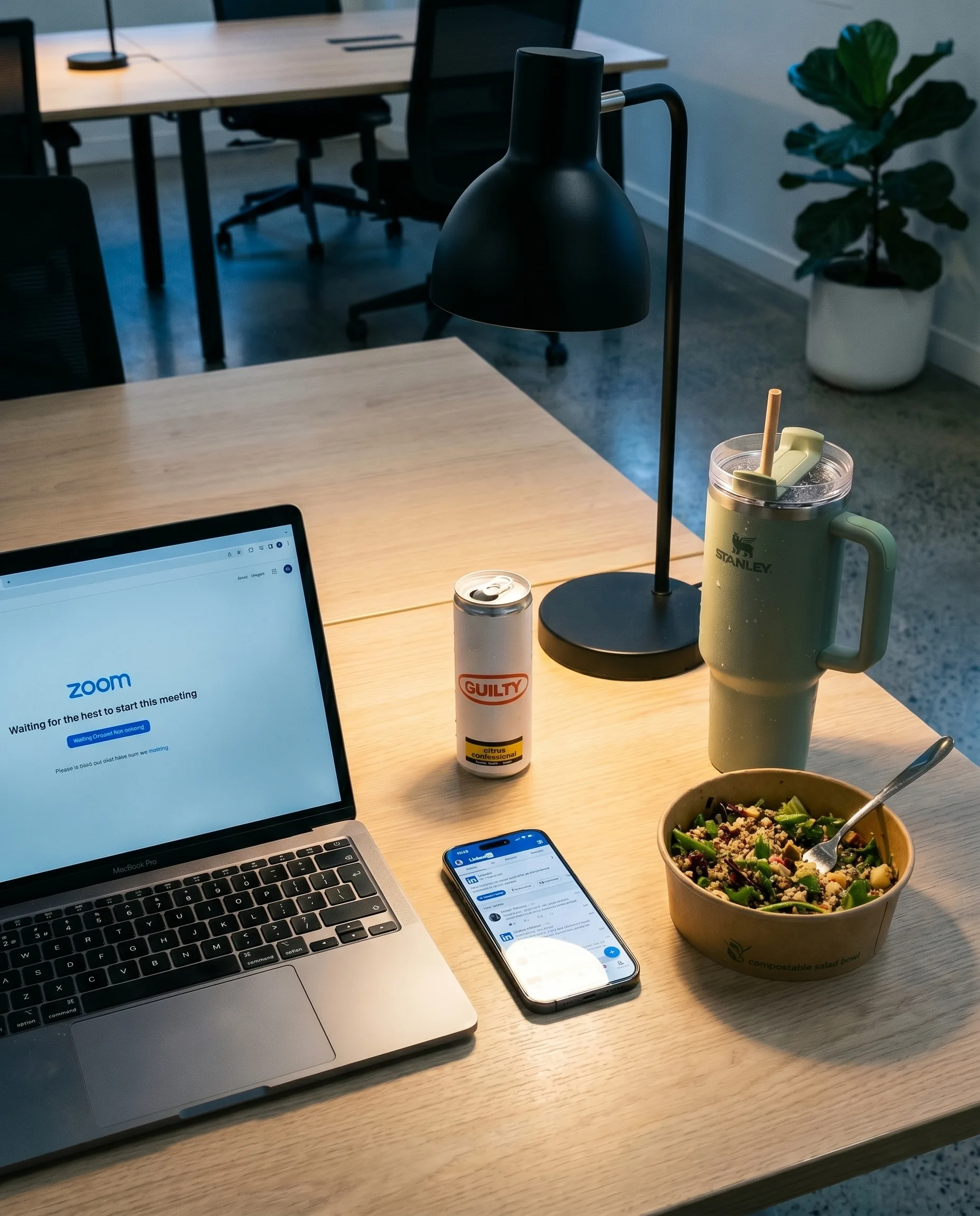 Forensic still life. Hot desk 27, Studio Society Surry Hills, late evening. MacBook in Zoom waiting room. iPhone showing LinkedIn. Sage Stanley Quencher. Half-eaten salad bowl. Citrus Confessional can. Empty office in background.